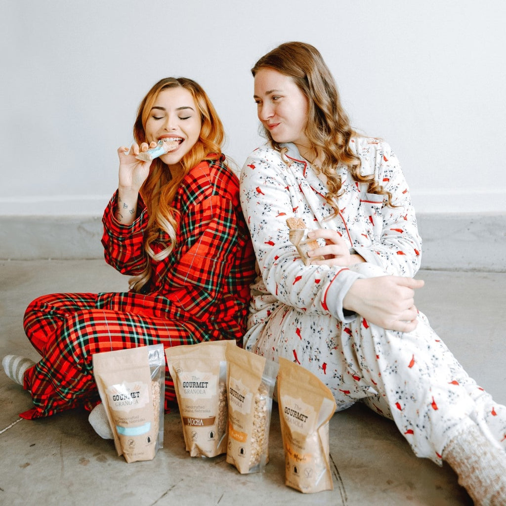 Two women enjoying granola bars with 4 bags of granola in front of them.
